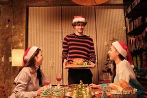 comida especial de la familia, el joven sirve pavo asado a los amigos, alegre con bebidas y disfruta comiendo, cena en el comedor de la casa decorado para el festival de navidad y la fiesta de las celebraciones de año nuevo. foto