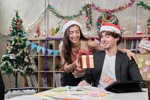 Sorprenda al equipo de dos compañeros de trabajo y al jefe dando regalos mientras el negocio trabaja antes de las vacaciones de la empresa en el lugar de trabajo de la oficina, está decorado para la celebración del festival de Navidad y la fiesta de año nuevo. foto