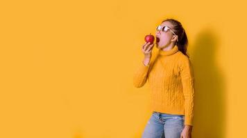 Cheerful young woman on yellow background in studio with a red apple in her hand she is opening her mouth to eat apples. The concept good health. Health lover photo