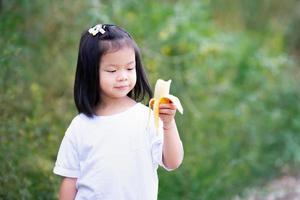 niño feliz disfruta comiendo plátano amarillo. dulce niña asiática sonriendo. foto