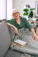 alegre mujer mayor sentada en el sofá leyendo un libro en casa. educación, maduro, concepto de ocio foto