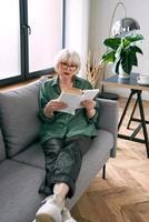 alegre mujer mayor sentada en el sofá leyendo un libro en casa. educación, maduro, concepto de ocio foto
