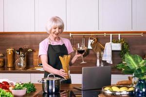 mujer alegre senior está cocinando en la cocina moderna por ordenador portátil. comida, educación, concepto de estilo de vida foto