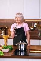 Mujer alegre senior está bebiendo vino tinto durante la cocción en la cocina moderna. comida, educación, concepto de estilo de vida foto