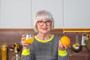 Cheerful pretty senior smiling woman in striped sweater  drinking orange juice while standing in the kitchen. Healthy, juicy lifestyle, home, senior people concept.