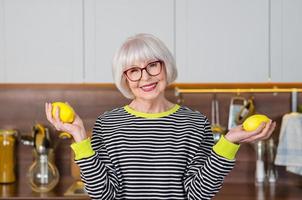 mujer sonriente bastante mayor alegre en suéter rayado que sostiene los limones para la limonada mientras está de pie en la cocina. estilo de vida saludable y jugoso, hogar, concepto de personas mayores. foto