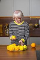 Cheerful pretty senior smiling woman in striped sweater holding lemons for lemonade while standing in the kitchen. Healthy, juicy lifestyle, home, senior people concept.