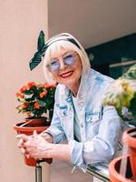 senior stylish woman with gray hair and in blue glasses and denim standing on balcony outdoors. photo