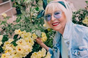 senior stylish woman with gray hair and in blue glasses and denim standing on balcony outdoors. photo