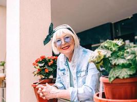 senior stylish woman with gray hair and in blue glasses and denim standing on balcony outdoors. photo