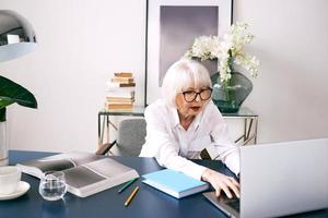Mujer de cabello gris hermosa senior cansada en blusa blanca trabajando en equipo portátil en la oficina. trabajo, personas mayores, problemas, encontrar una solución, concepto de experiencia foto