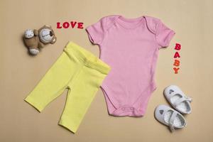 vista superior de primer plano. Maqueta de mono rosa en blanco, pantalón amarillo y sandalias blancas para recién nacidos sobre un fondo beige, con espacio de copia: plantilla de maqueta de ropa de bebé perfecta foto