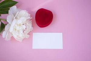 Close up of a greeting card mockup. On a pink background, a business card, a red jewelry box and a white peony. Top view, flat layout. photo