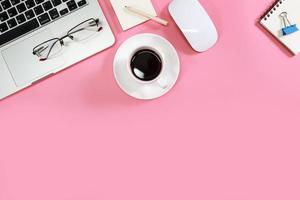 Mesa de espacio de trabajo plano con computadora portátil, suministros de oficina, taza de café, teléfono celular, tableta y taza de café sobre fondo rosa foto