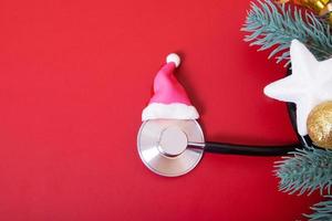 Estetoscopio médico laico plano en gorro de Papá Noel y adornos navideños sobre fondo rojo. concepto médico de navidad foto