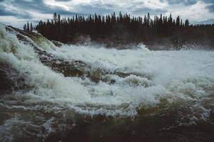 Waves and splashes of mountain river on background of forest and dramatic sky. Forest river water landscape photo