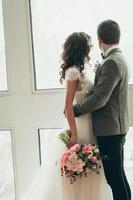 La novia y el novio con ramo de novia de pie junto a la ventana grande, vista posterior