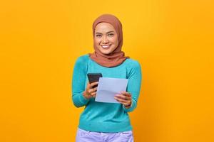 Feliz joven mujer asiática sosteniendo smartphone y factura aislado sobre fondo amarillo