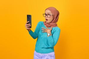 Conmocionada joven y bella mujer asiática mira la pantalla del teléfono inteligente aislada sobre fondo amarillo foto