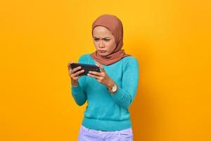 Conmocionada joven asiática jugando un videojuego en un teléfono inteligente sobre fondo amarillo
