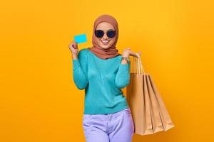 Smiling young Asian woman holding shopping bags and credit card on yellow background photo