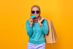 Cheerful young Asian woman biting credit card and hold shopping bags on yellow background photo