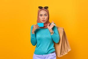 Shocked young Asian woman holding credit card and shopping bags on yellow background photo
