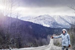 Mujer haciendo autostop en una carretera de invierno con hermosas montañas nevadas en segundo plano.