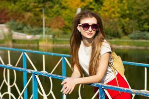Beautiful girl in the park with a fashionable backpack and in sunglasses on vacation leaned on the handrail photo