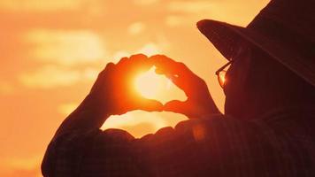 silhouet van senior boer in een hoed die bij zonsondergang in de verte kijkt en een hartvorm maakt op een gouden hemelachtergrond. video