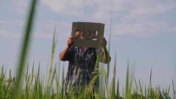 A senior farmer standing in a rice field at sunset and holding a New Year 2022 sign.