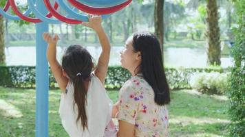 La madre ayuda a su pequeña hija a colgarse de las barras de colores en el patio de recreo. jugar es aprender en la niñez. video