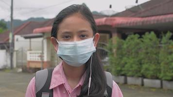 portret middelbare school meisje met rugzak met gezichtsmasker en koptelefoon kijken naar de camera. video