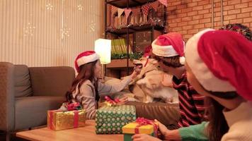 kvinnlig vän av asiatisk etnicitet retar hund med röd hatt, medan förbereder presenter för julaftonsfirande i hemmets vardagsrum, vackert dekorerad, semesterfestival och gott nytt år.