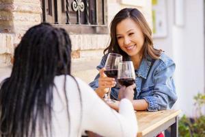 Two women making a toast with red wine sitting at a table outside a bar.