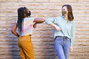 Multiethnic young women wearing masks greeting at each other with their elbows photo