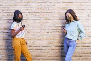 Mujeres jóvenes multiétnicas con máscaras que utilizan teléfonos inteligentes separados para respetar la distancia social.