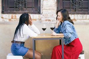 dos amigos multiétnicos hablando sentados en una mesa fuera de un bar mientras beben una copa de vino tinto. foto