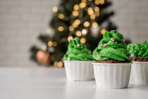 Cupcakes con forma de árbol de Navidad, rodeados de adornos festivos y luces de fondo