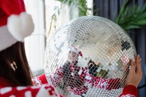 Young woman in santa hat holding mirror disco ball standing by the Christmas tree photo