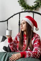 Young woman in red sweater and santa hat sitting on the bed celebrating christmas having cup of hot drink photo