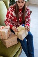 Mujer joven en suéter rojo envolver un regalo sentado en el sofá foto