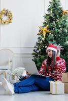 Mujer joven sentada cerca del árbol de Navidad en una sala de estar decorada con chat en línea en la computadora portátil