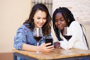 Two women looking at their smartphone together while having a glass of wine. photo