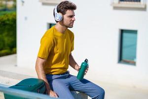 Male sitting outside using an aluminum water bottle, headphones and backpack. photo