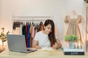 Mujer asiática emprendedora de tienda de ropa trabajando y tomando nota en el portátil con el portátil en la mesa. foto
