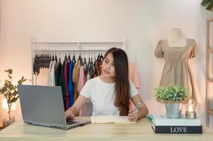 Mujer asiática emprendedora de tienda de ropa con portátil y tomando nota en el portátil en el escritorio