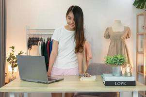 Mujer asiática emprendedora de la tienda de ropa online de pie usando un portátil y comprobando el inventario en el escritorio