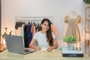 Mujer asiática emprendedora de tienda de ropa trabajando y tomando nota en el portátil con el portátil en la mesa. foto