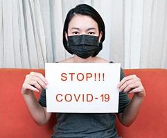 Woman wearing health mask and holding paper with text stop covid 19 photo